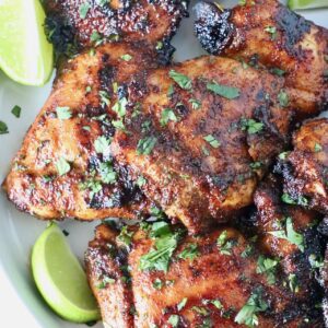 grilled chicken thighs stacked up on a plate with fresh cilantro and lime wedges
