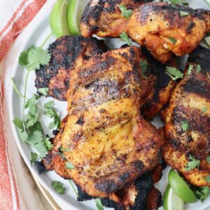 grilled chicken thighs stacked up on a plate with fresh cilantro and lime wedges
